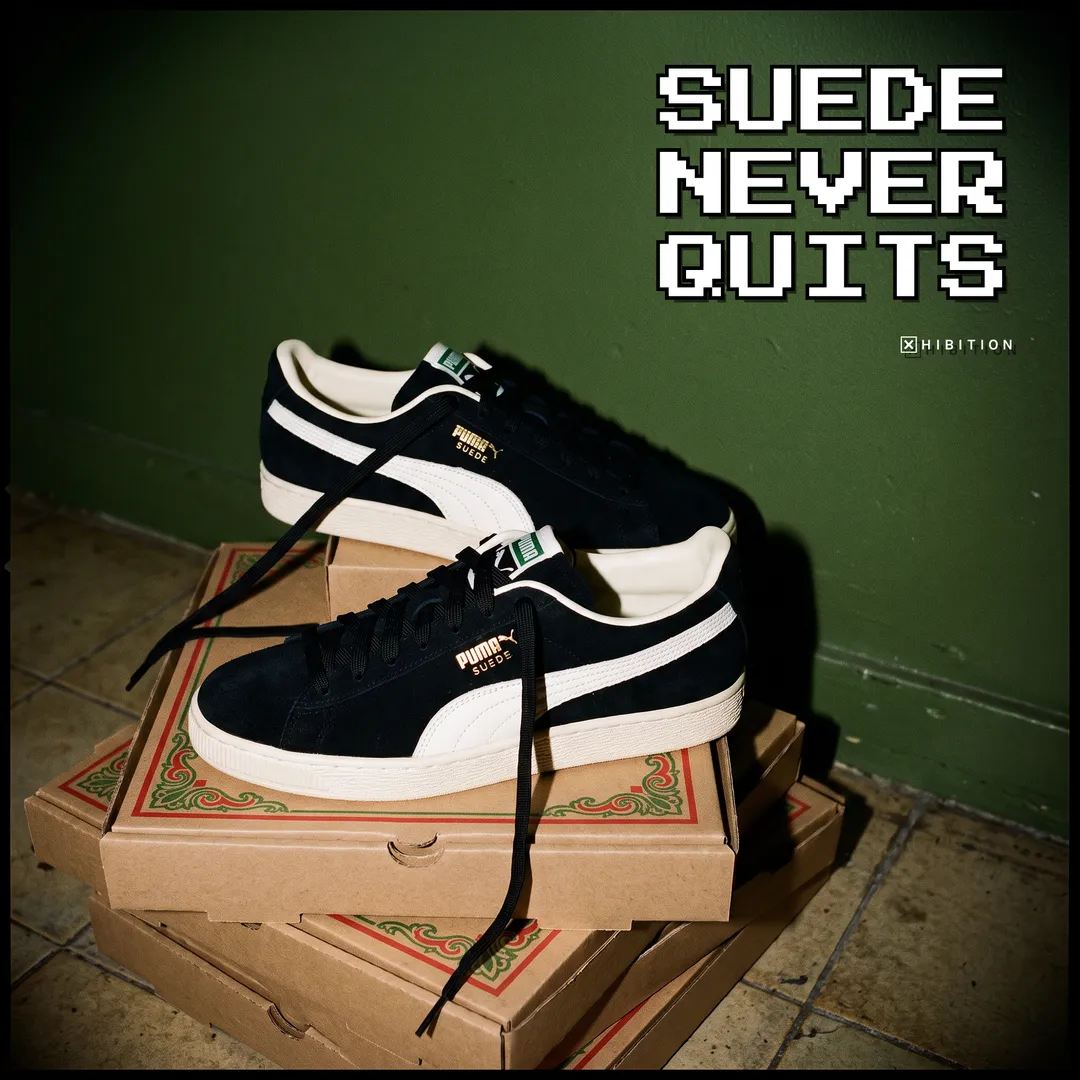 A pair of PUMA Suede Charles F. Stead I sneakers stacked on cardboard shoe boxes against a deep green wall, showing the stitched white formstrip and the gold PUMA Suede side stamp.